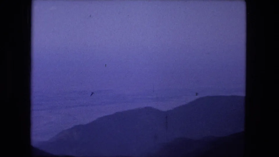 Cataloged 1976 8mm frame showing hazy blue mountain silhouettes and a dark foreground ridge with visible film dust and soft exposure.