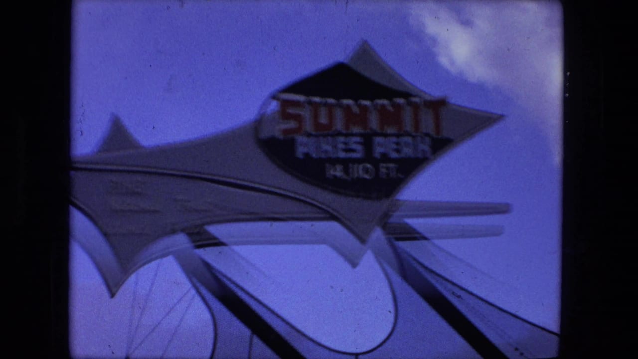 Closer 1976 8mm view of the Summit Pikes Peak sign at 14,110 ft, with blue sky, clouds, and structural supports visible.