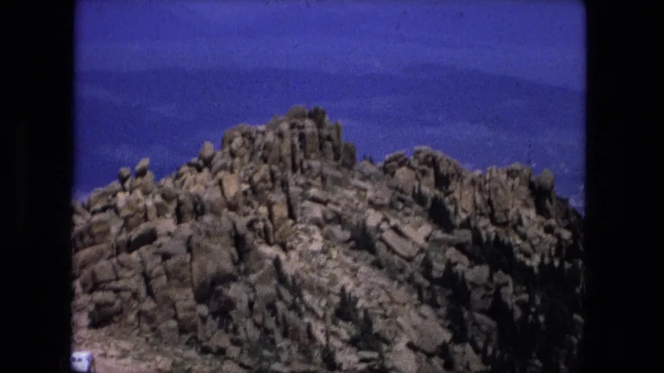 1976 8mm frame of rugged rocky summit terrain with sparse trees, hazy lower land, and blue sky in the distance.