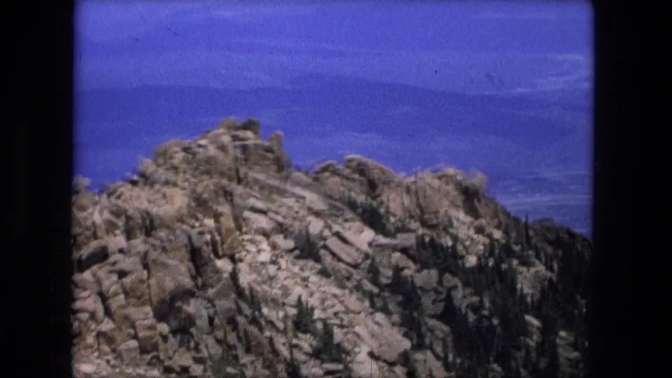 1976 8mm travel frame showing a rocky mountain ridge, scattered trees, and a hazy distant landscape under blue sky.