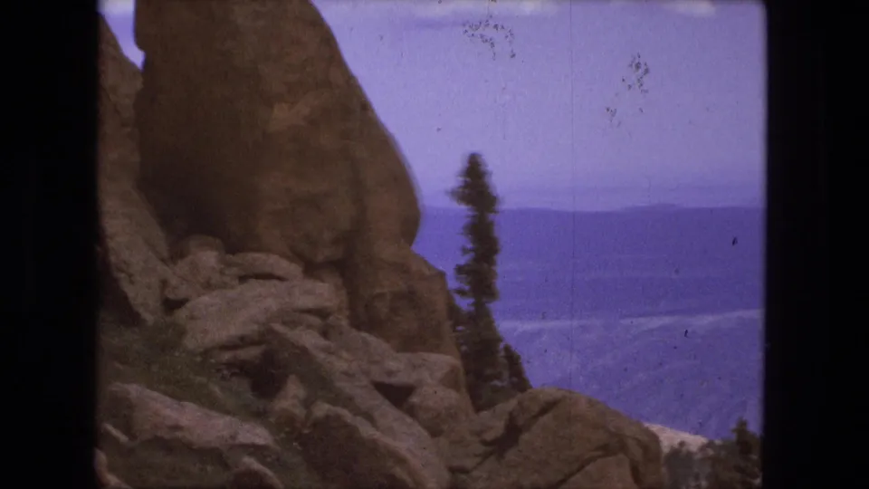 1976 8mm frame showing a rocky overlook with evergreen trees and a distant blue valley beyond.