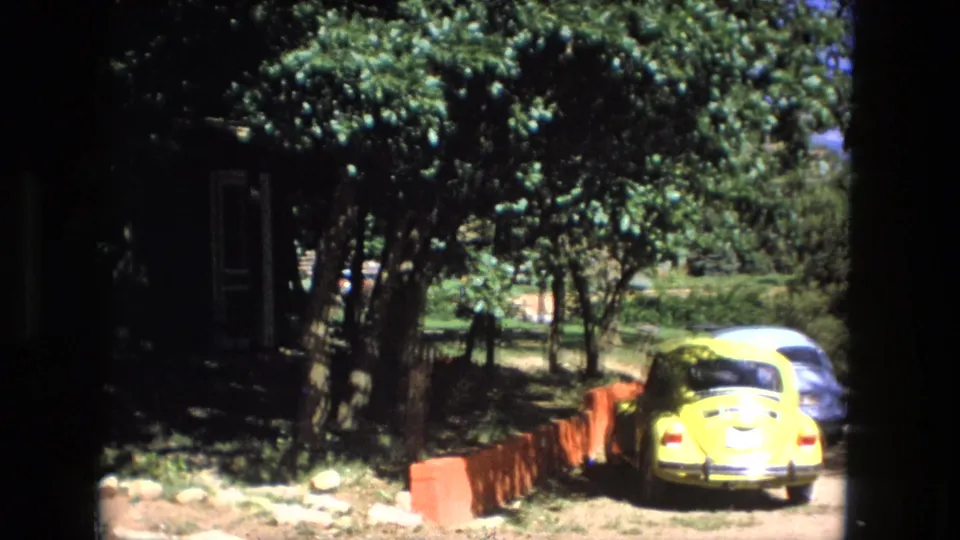 Fort Wayne 1976 driveway scene with a yellow compact car, a pale car behind it, trees, red edging, and a shaded building.
