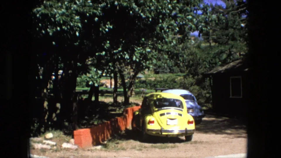 1976 Fort Wayne exterior with a yellow compact car on a gravel driveway, another car behind it, trees, red border, and a small building.