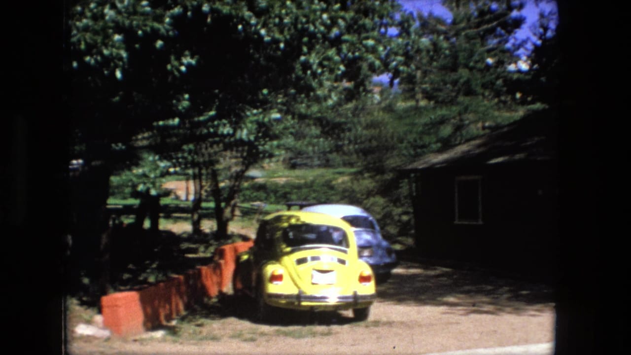 Yellow compact car parked near trees and a small dark building beside a dirt road.