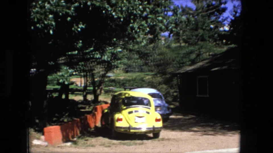 1976 Fort Wayne home-movie frame showing a yellow compact car, trees, red roadside barrier, and small building in bright sun.