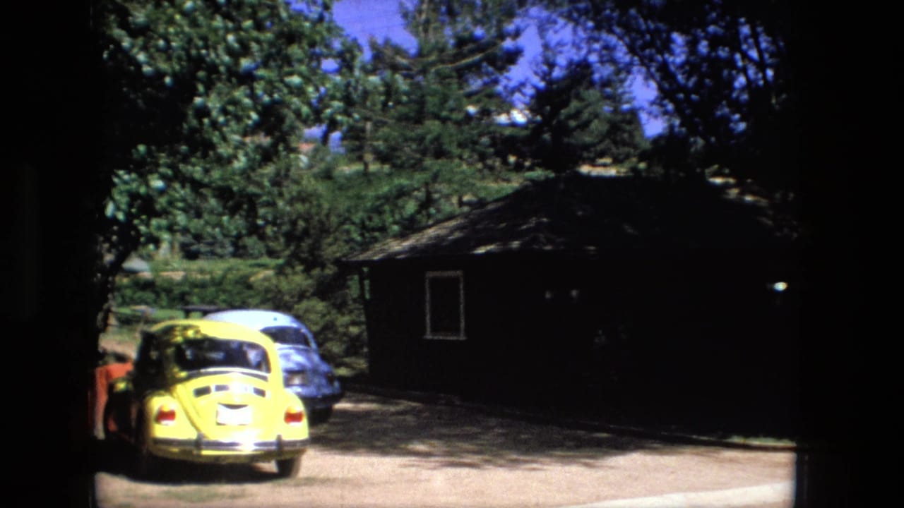 Fort Wayne 1976 frame with yellow car, pale parked vehicle, tree shade, and a small roadside building in Super 8 color.