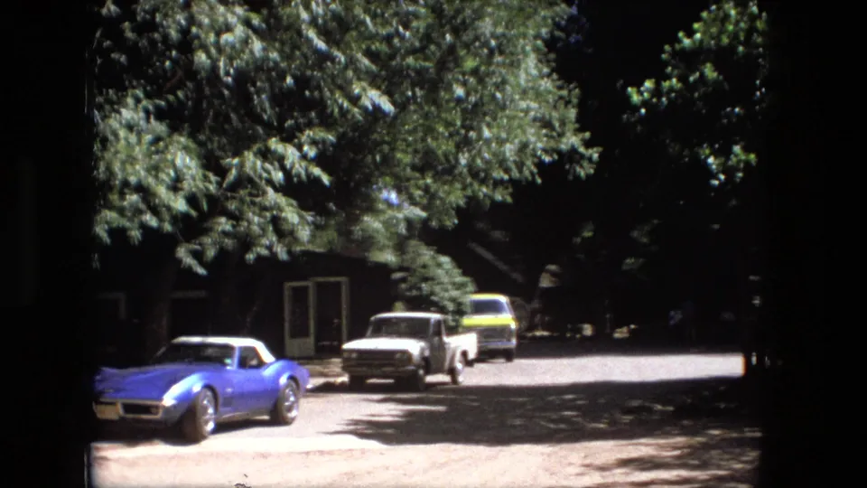 1976 Fort Wayne home-movie frame showing a blue sports car, white pickup, yellow vehicle, trees, and a shaded dirt road.