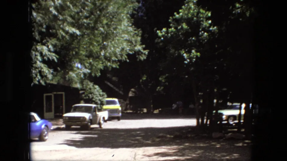 Fort Wayne 1976 frame with a white pickup, yellow vehicle, partial blue sports car, and shaded roadside trees.
