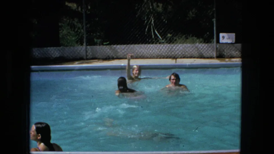1976 Fort Wayne home-movie frame showing swimmers spread across a blue pool beside a chain-link fence and 9 ft depth marker.