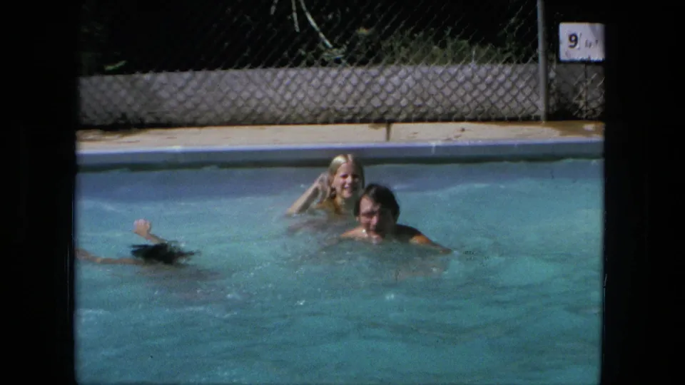 1976 Fort Wayne pool scene with a blonde woman and man smiling in the water while another swimmer crosses the frame.