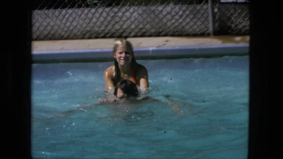 1976 Fort Wayne home-movie frame of a blonde woman and man playing in a swimming pool with soft 8mm grain.