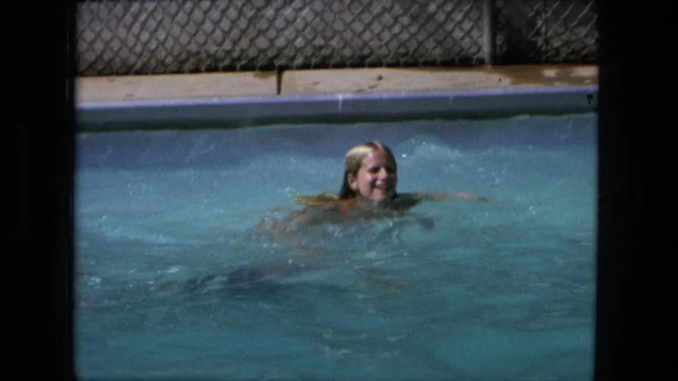 1976 Fort Wayne frame showing a blonde woman swimming through blue pool water with the pool edge and fence behind her.
