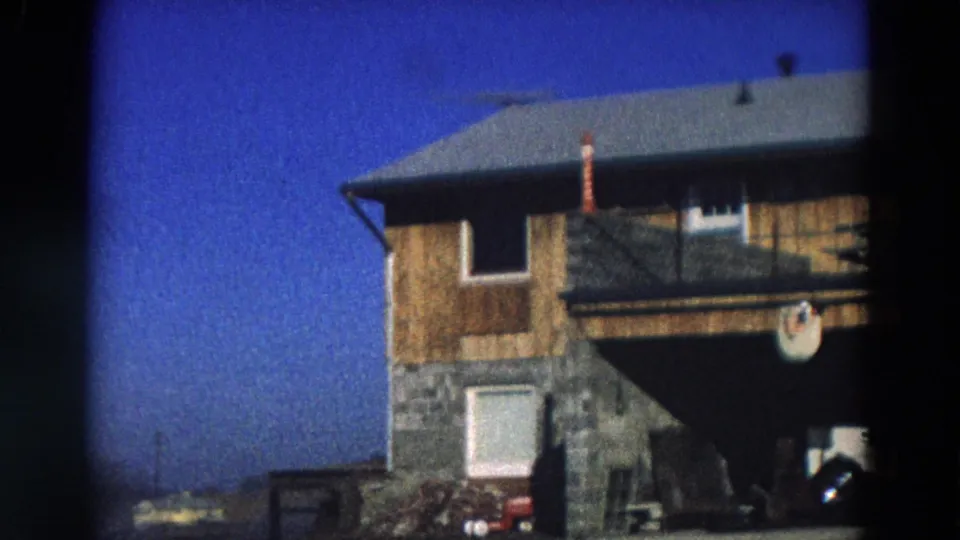 1968 Diller, Nebraska 8mm frame showing the house corner, white door, stone base, porch rail, and a partial vehicle below.