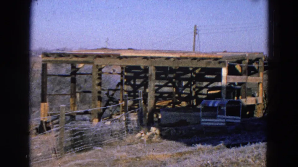 1968 Diller, Nebraska 8mm frame of a wooden shelter structure with wire fencing, dry grass, and a small vehicle in daylight.