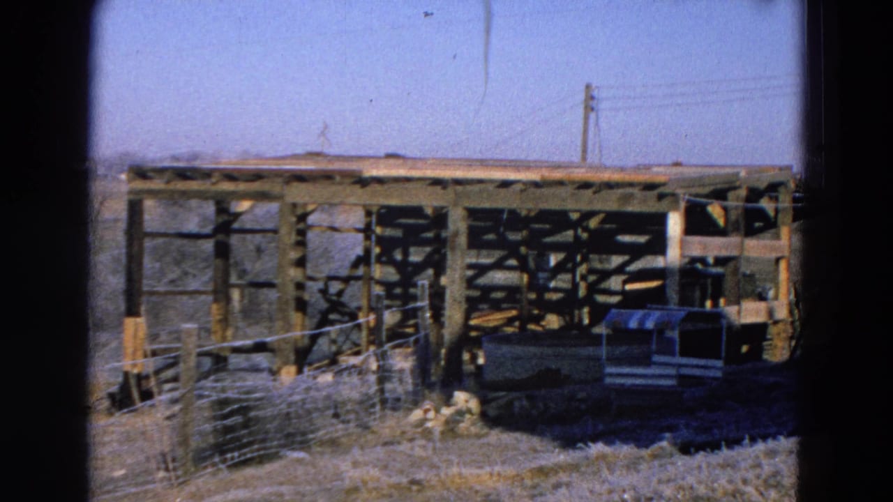 Wooden barn frame with wire fencing, animal shelter, utility pole, power lines, dry grass, and pale sky.