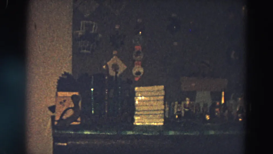 Home-movie view of the garage-party bar area in Diller, Nebraska, with bottles, stacked items, and dim wall decor.