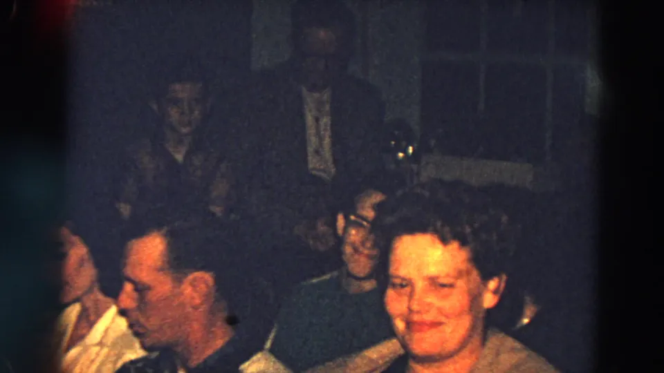 1968 Diller, Nebraska garage-party frame showing adults mingling, including a smiling woman close to the camera.
