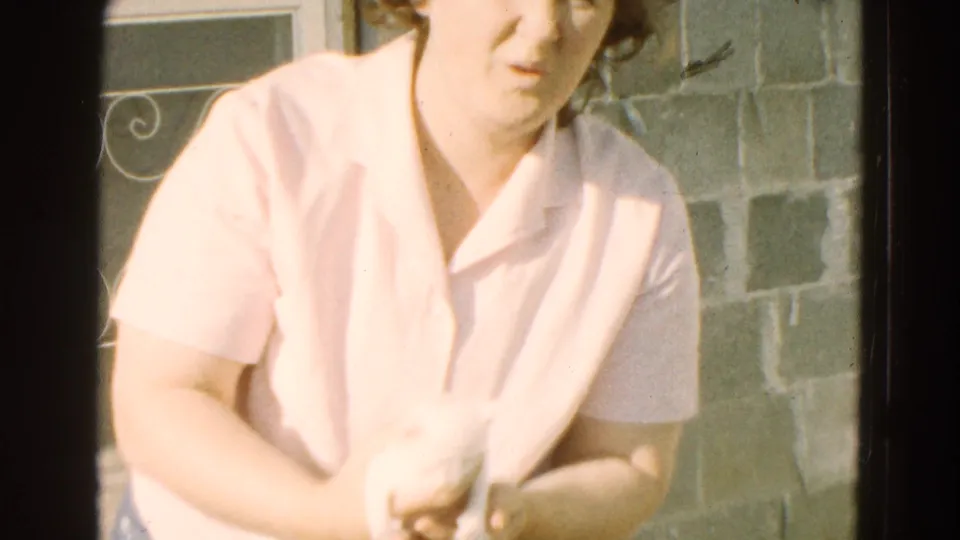 An adult woman in a pale collared shirt holds a very young puppy in bright outdoor light, with masonry visible behind her.