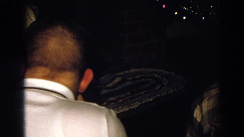 1968 Diller, Nebraska 8mm frame showing the back of a seated man near a brick wall, patterned textile, and distant lights.