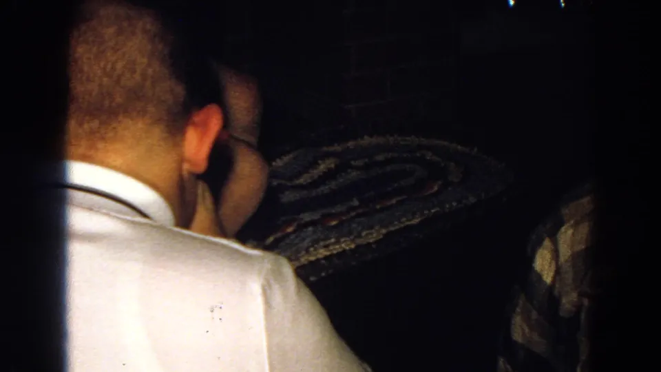 1968 Diller, Nebraska frame showing a man in a white shirt from behind, beside a patterned rug or textile and partial nearby person.