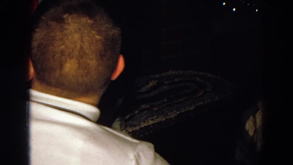The man remains turned away in a dim Diller, Nebraska interior, with a patterned rug or textile visible beyond him.