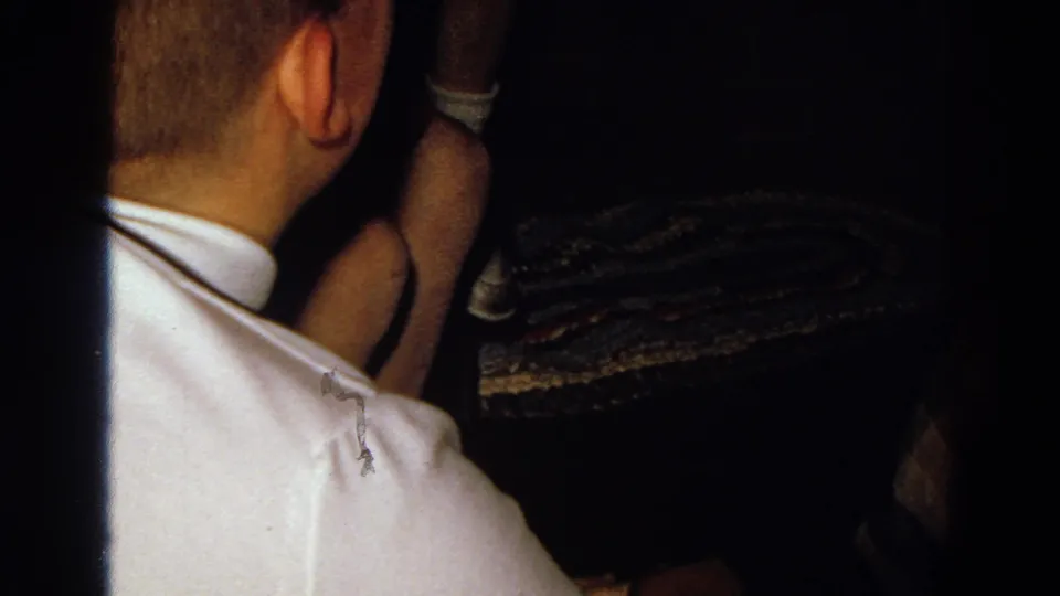1968 Diller, Nebraska 8mm frame showing a person from behind in a white shirt, seated close to others in a low-light interior.