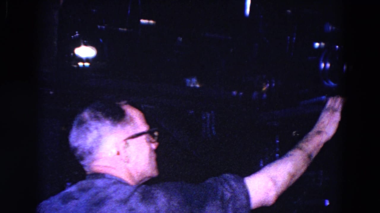 Man in glasses reaches toward dark factory machinery beside large paper rolls.