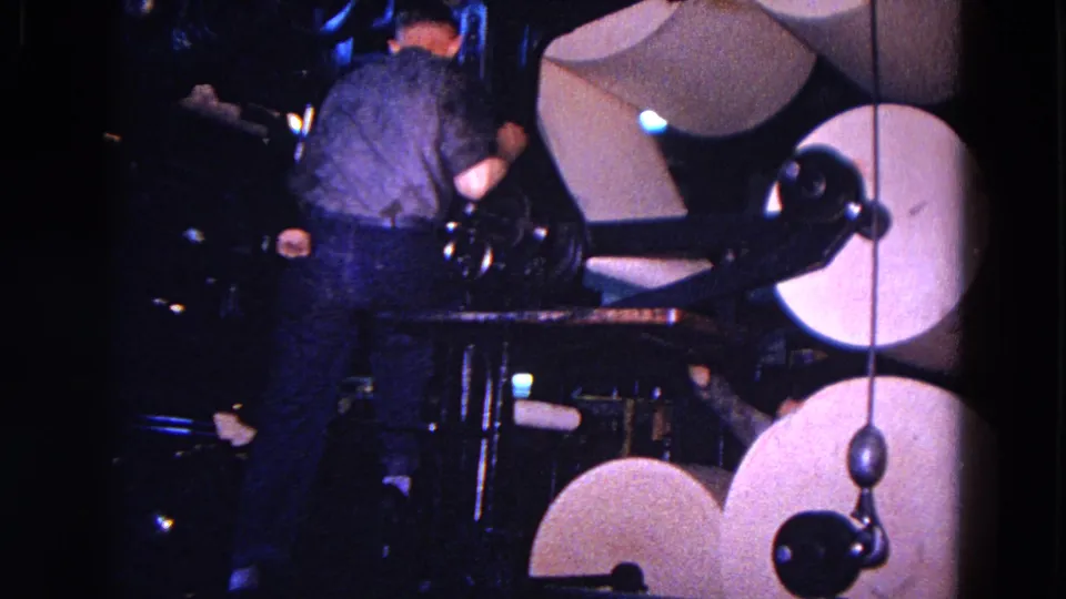 A worker stands close to large paper rolls and mechanical arms in the dim 1967 Trenton factory scene.