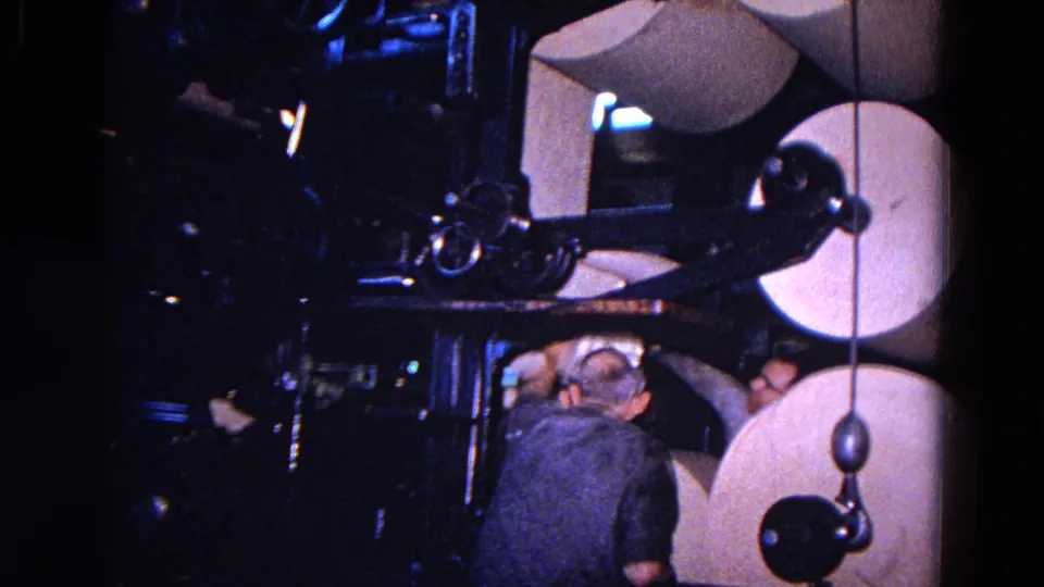 A worker crouches low beside large paper rolls and mechanical parts in the 1967 Trenton factory footage.