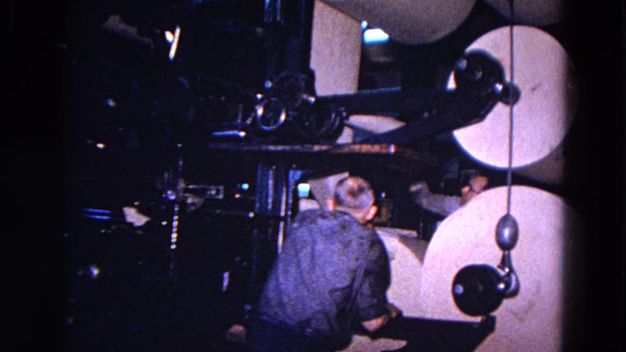 Man in a blue shirt works beside large paper rolls and dark machinery.