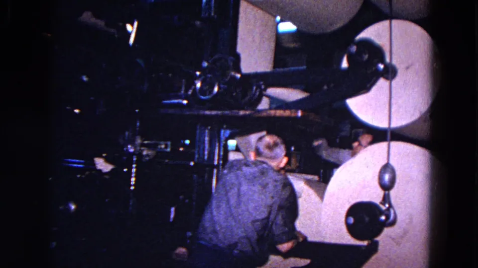 In 1967 Trenton, a man in a blue shirt and glasses stands near large paper rolls inside a dim industrial machine area.
