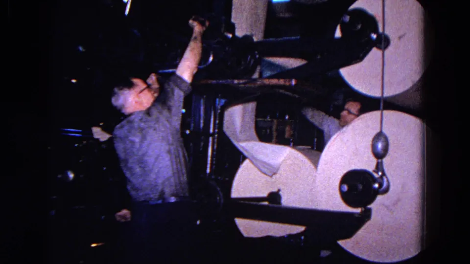 The worker reaches up toward an overhead part of the paper machine, with large rolls and dark factory equipment around him.