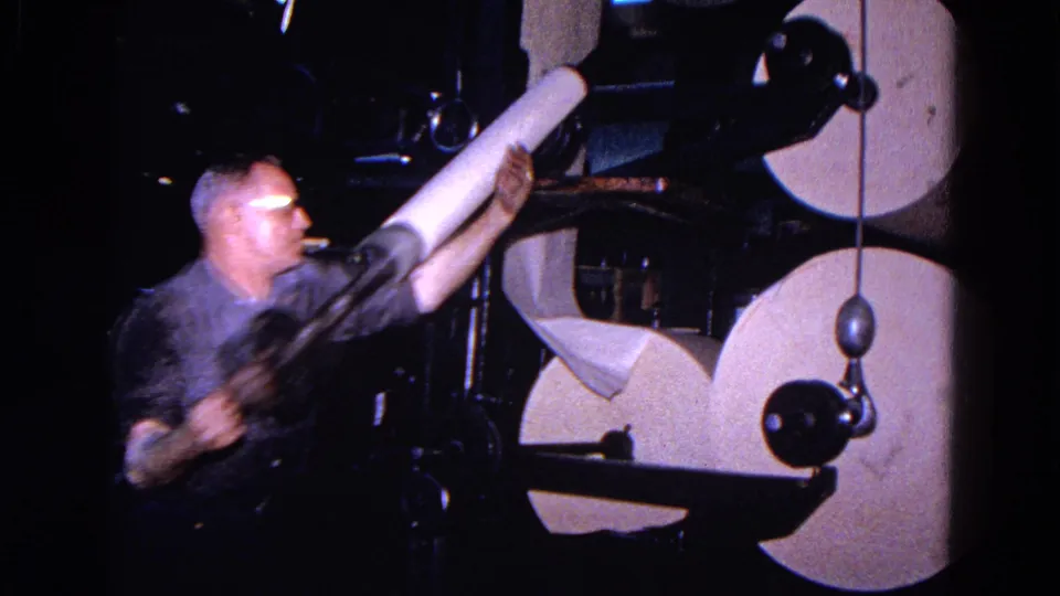 A man in glasses carries or removes a white cylindrical spool near the paper mill machine in the dim factory setting.