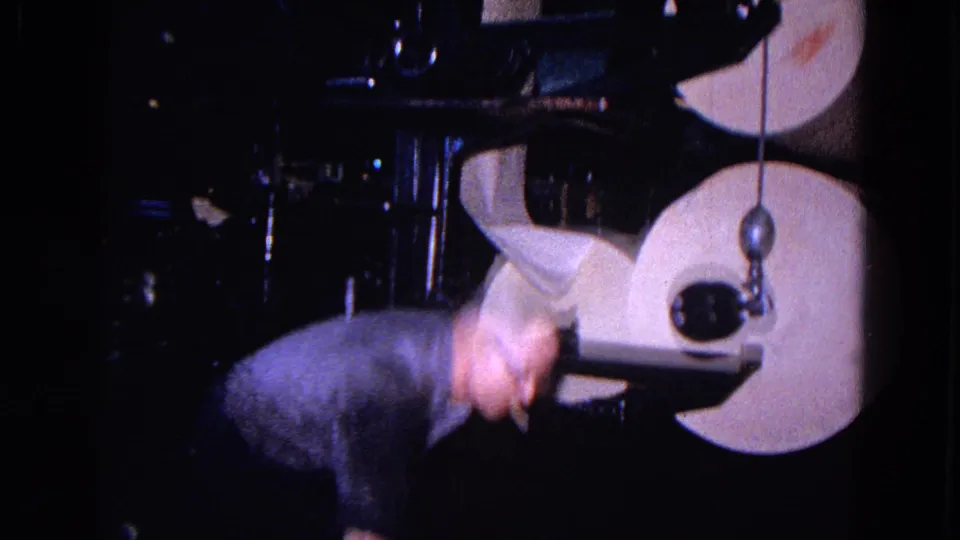 The worker bends down beside the large paper rolls and dark machine frame, partly blurred by movement in the 8mm footage.