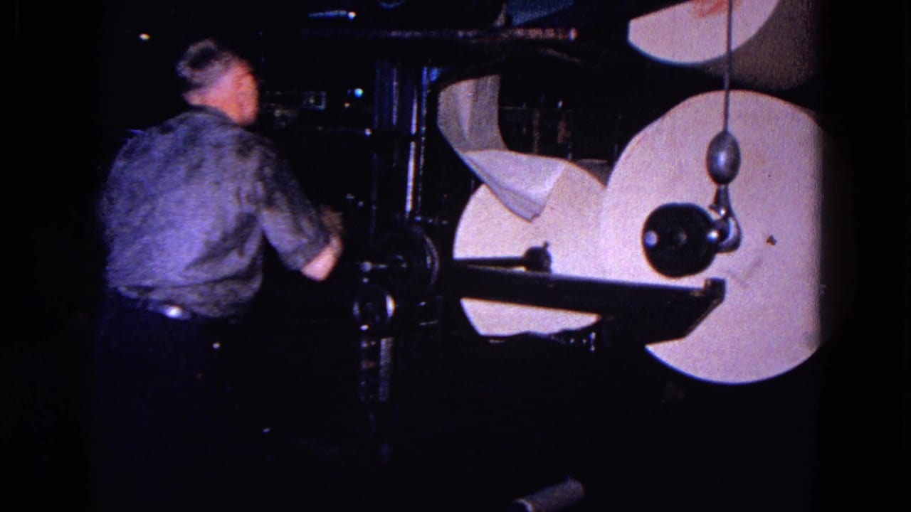 Man stands beside large white paper rolls on a dark industrial machine.