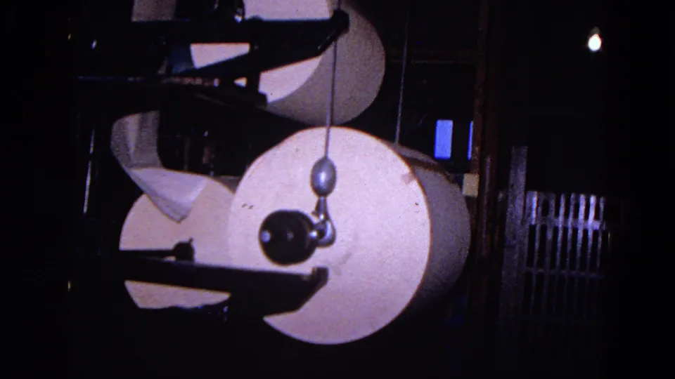 Large rolls of white paper are framed by black industrial press components, with a hanging weight visible in the foreground.