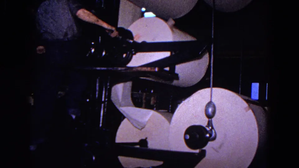A worker in a dark shirt reaches into a large paper roll machine, with multiple white rolls and press arms visible around him.