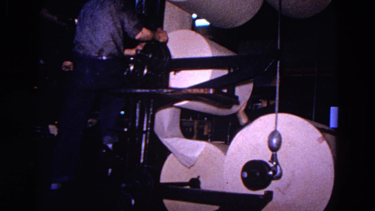 In 1967 Trenton footage, a man stands close to large paper rolls and dark metal press machinery in dim light.