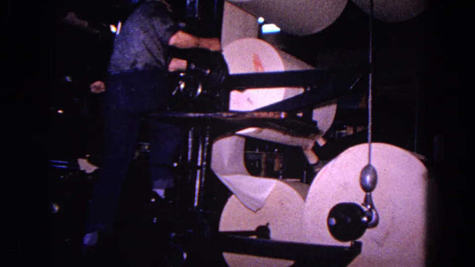 The worker leans into the printing press area as large paper rolls and black metal arms fill the frame.