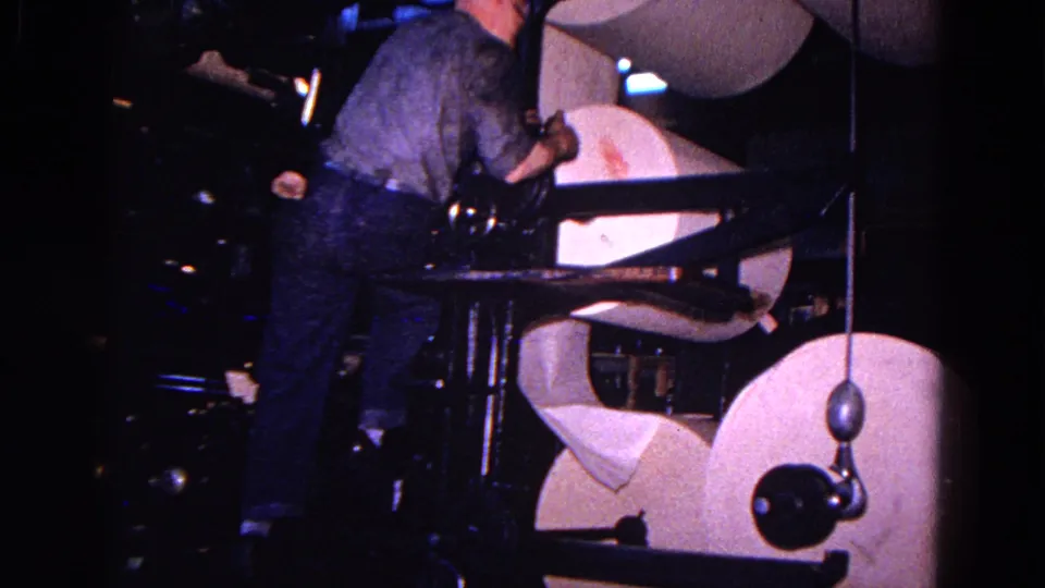 Seen from behind, the man works at the press near pale paper rolls and heavy dark machine parts.
