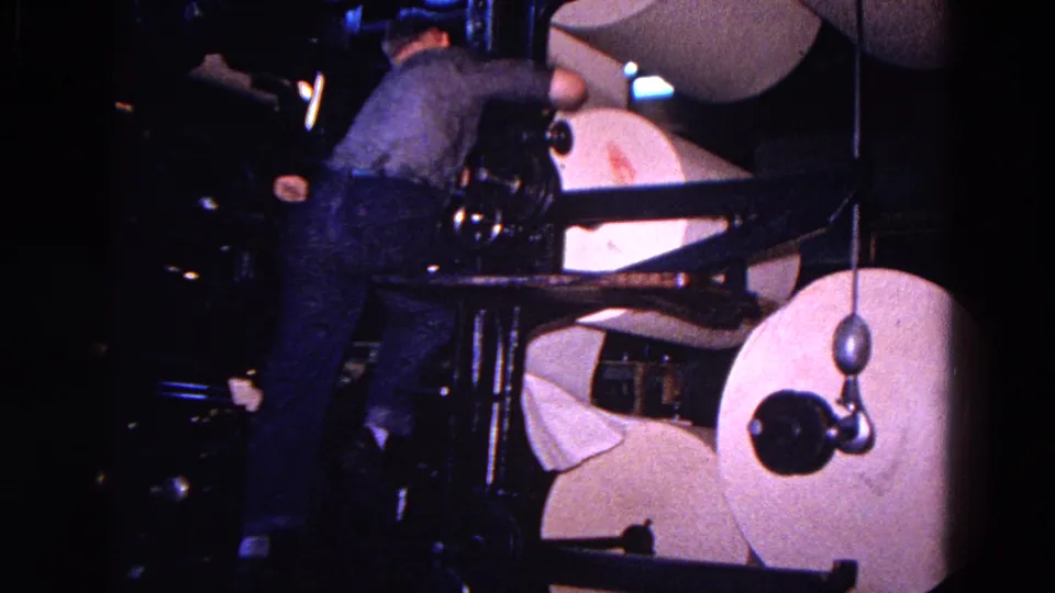 The worker bends toward the press mechanism, with a large round paper roll prominent in the foreground.