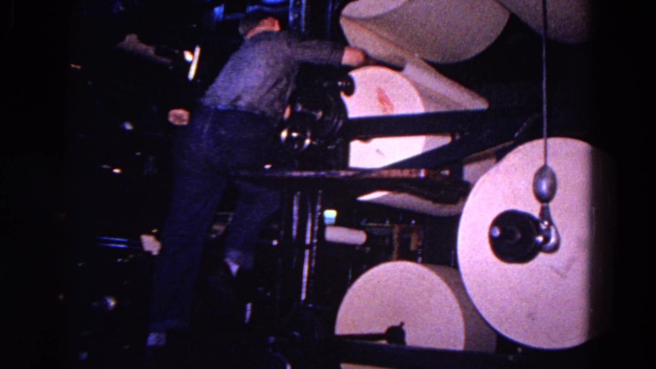 In supplied 1967 Trenton footage, a man works near large paper rolls and black press arms inside a dim industrial printing space.