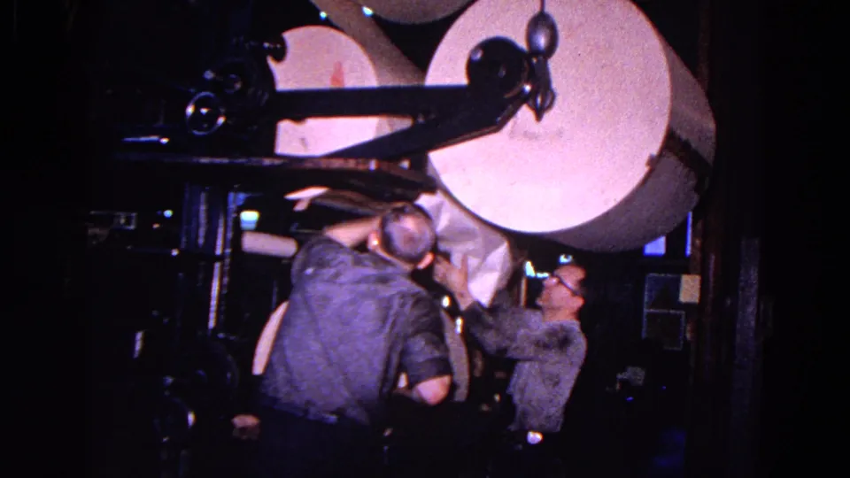 Two adult men appear to guide paper through a large printing press, with a pale roll overhead and machinery surrounding them.