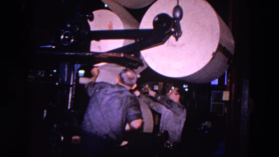 In the final frame, two men work closely under a large paper roll on an industrial printing press in the supplied Trenton 1967 footage.
