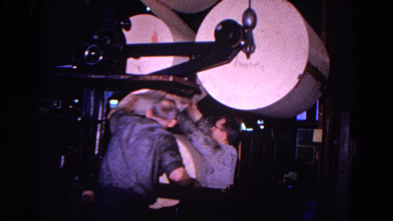 Two workers stand beside large white paper rolls on a dark industrial machine.