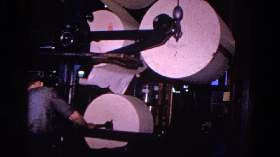 A worker in the Trenton industrial facility appears to feed or adjust paper beneath a large roll on the machine.