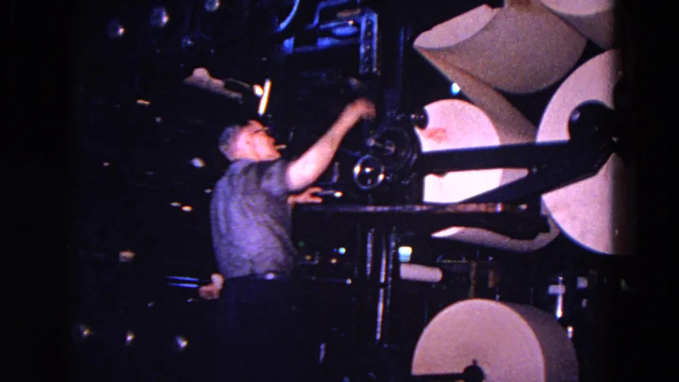 A worker reaches toward the machinery controls near several large paper rolls in the Trenton industrial scene.