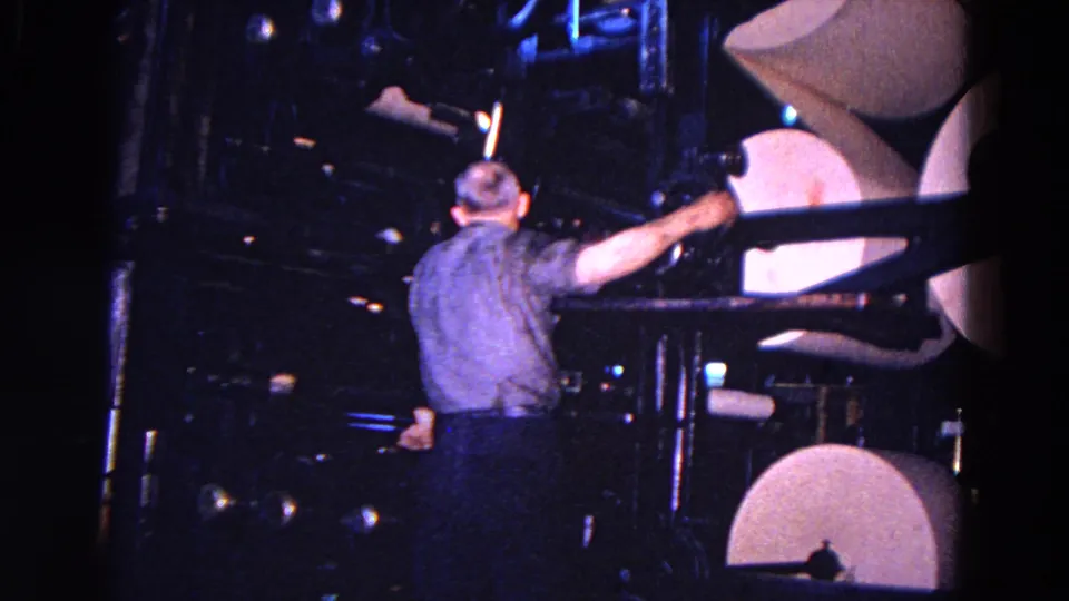 Rear view of a 1967 Trenton factory worker reaching across dark machinery toward large rolls of paper.