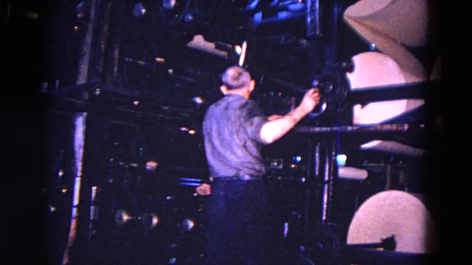 Factory worker in Trenton, New Jersey, turns a round control beside large paper rolls in 1967 8mm footage.