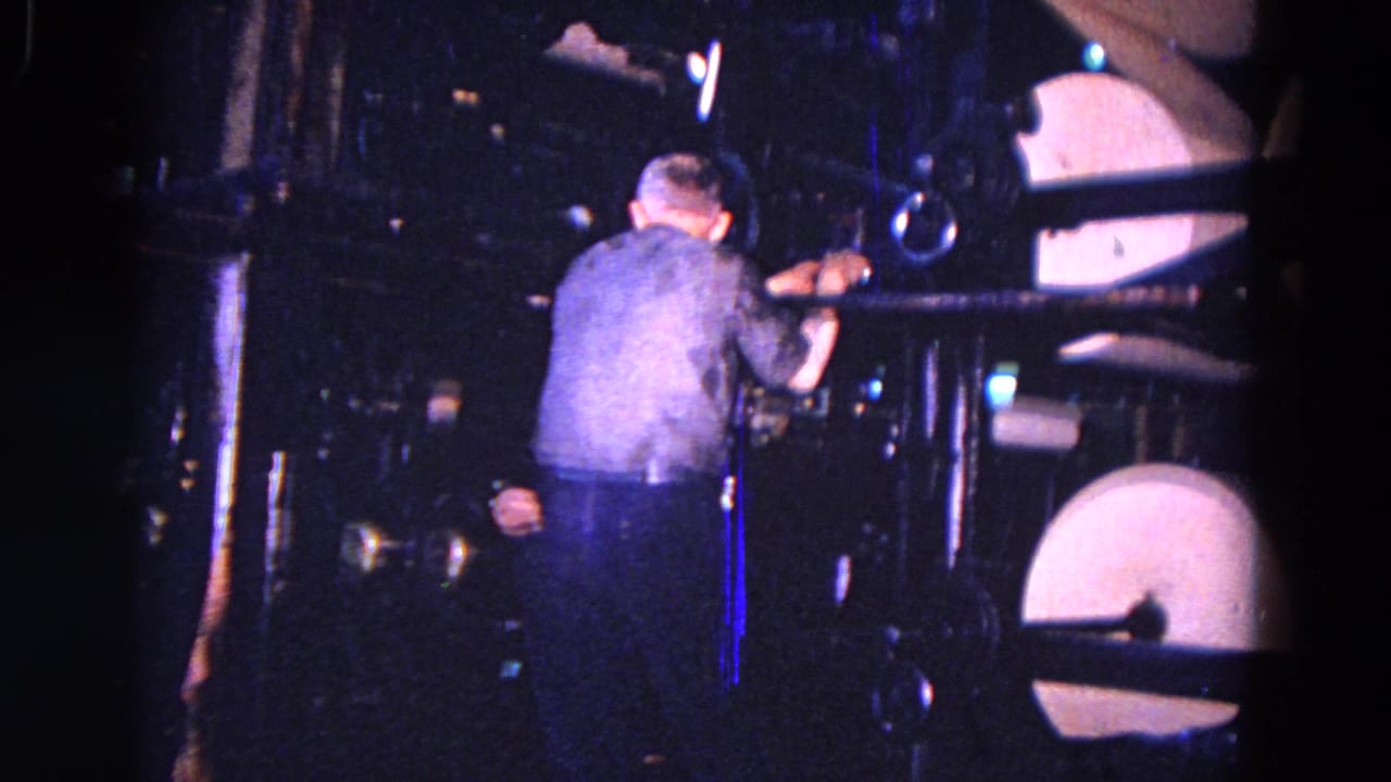 In 1967 Trenton, New Jersey metadata context, a worker appears beside heavy machinery with pale round drum-like parts in a dark factory space.